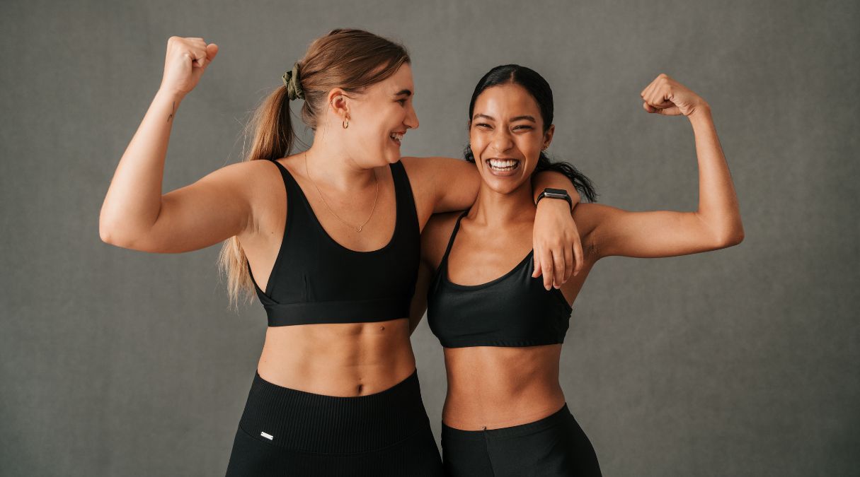 Two women in athletic wear smiling and flexing their arms, celebrating fitness