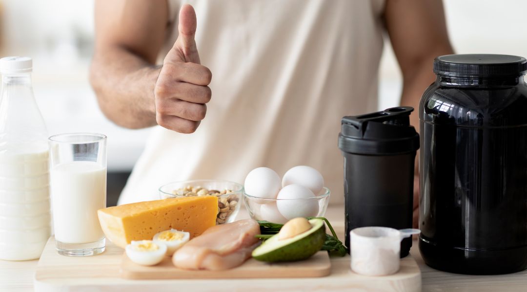 Man giving a thumbs-up next to a table featuring protein-rich foods, including eggs, cheese, chicken, and a protein shake