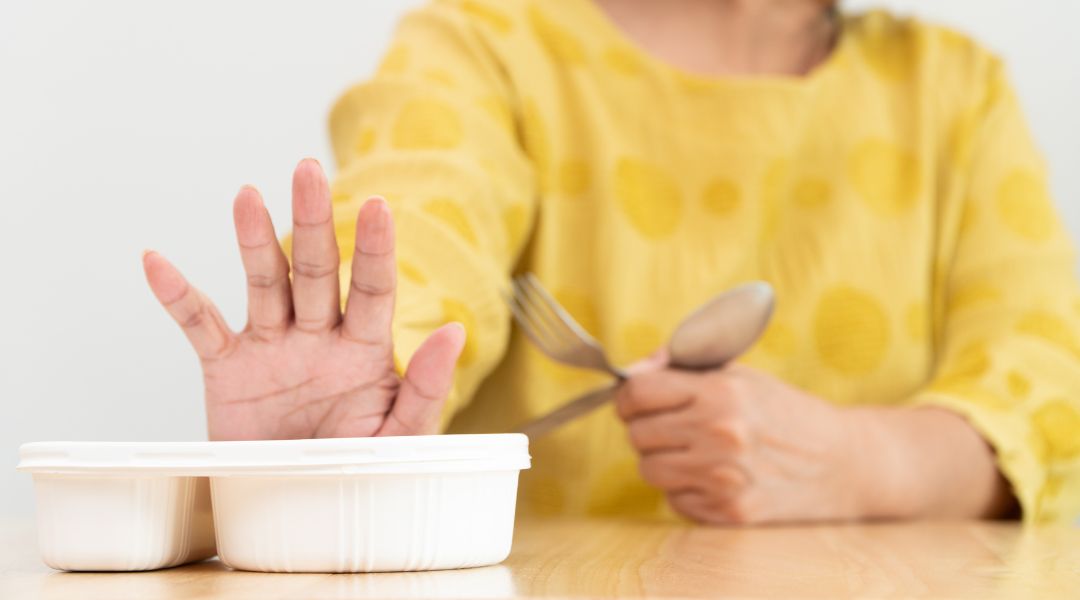 A woman in a yellow shirt raises her hand to decline food from a white container, holding cutlery in her other hand