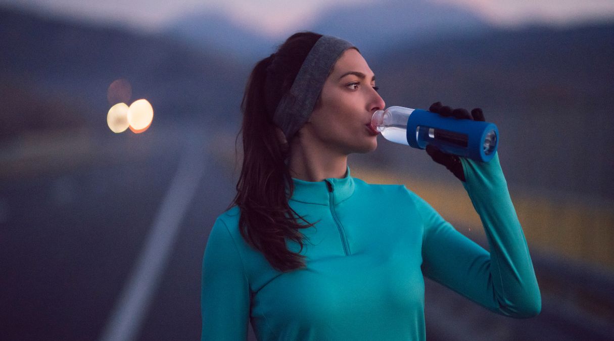 Woman drinking from a bottle while exercising outdoors at dusk, wearing a turquoise jacket