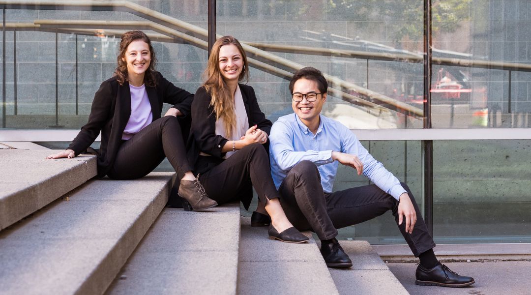 Three dietitians in business casual attire sitting on outdoor steps, smiling at the camera