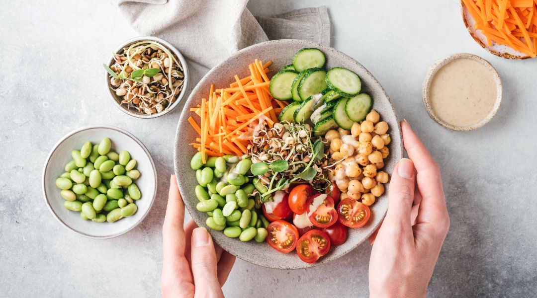 Plate of colorful vegetables and legumes, including carrots, cucumbers, tomatoes, chickpeas, and edamame, with small bowls of toppings nearby