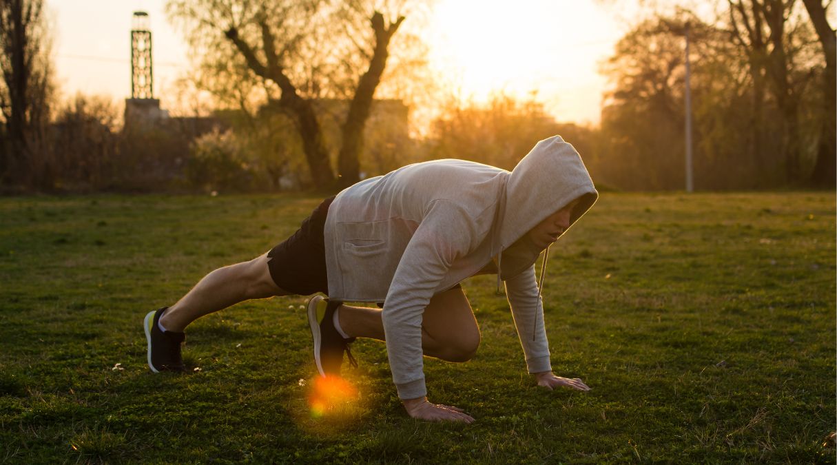 Man in a hoodie doing mountain climbers exercise on grass at sunrise