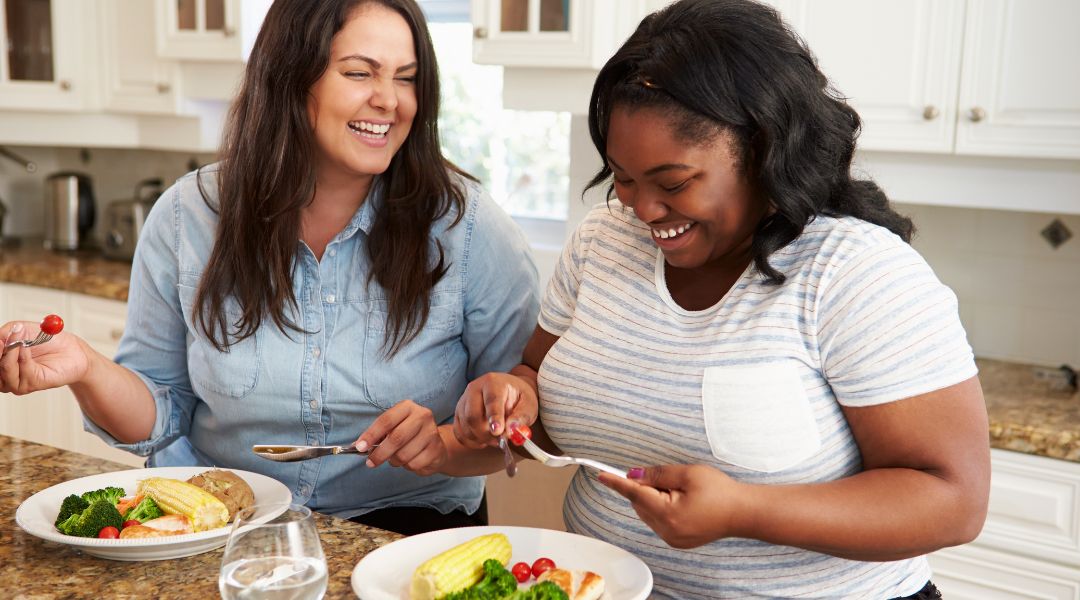 Two women laughing while eating a healthy meal in a kitchen, enjoying each other’s company