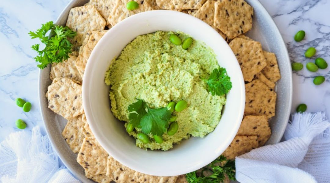 A bowl of green edamame hummus garnished with parsley, surrounded by seed crackers