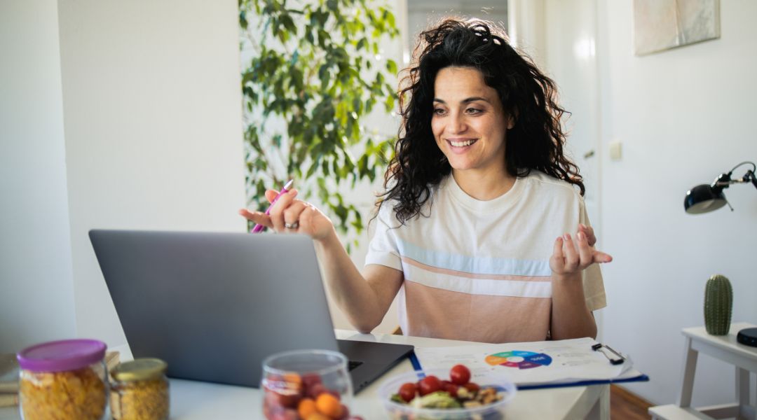 A smiling woman gesticulates while working on a laptop at home, surrounded by graphs and healthy snacks