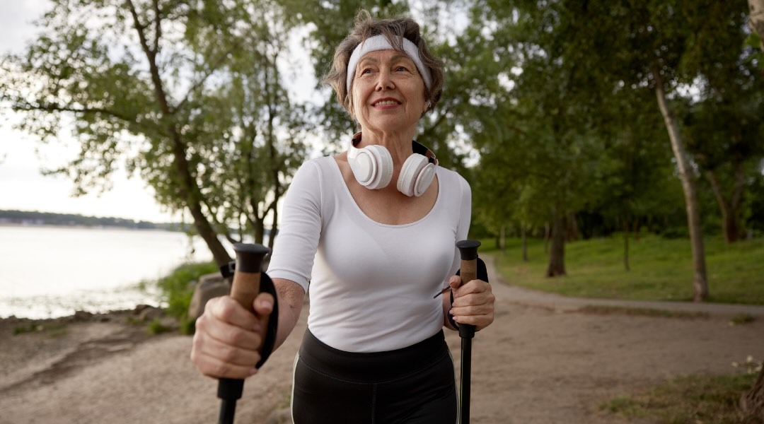 An elderly woman walks with Nordic walking poles in a natural setting, smiling confidently and wearing headphones.