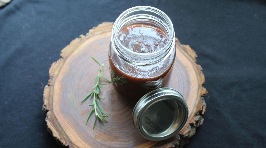 Mason jar of dark barbecue sauce with a sprig of rosemary on a rustic wooden surface, lid resting beside it