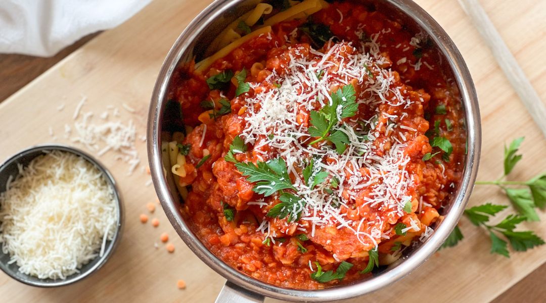 Pot of pasta with rich red bolognaise sauce, topped with grated Parmesan and fresh parsley, with extra cheese in a small bowl