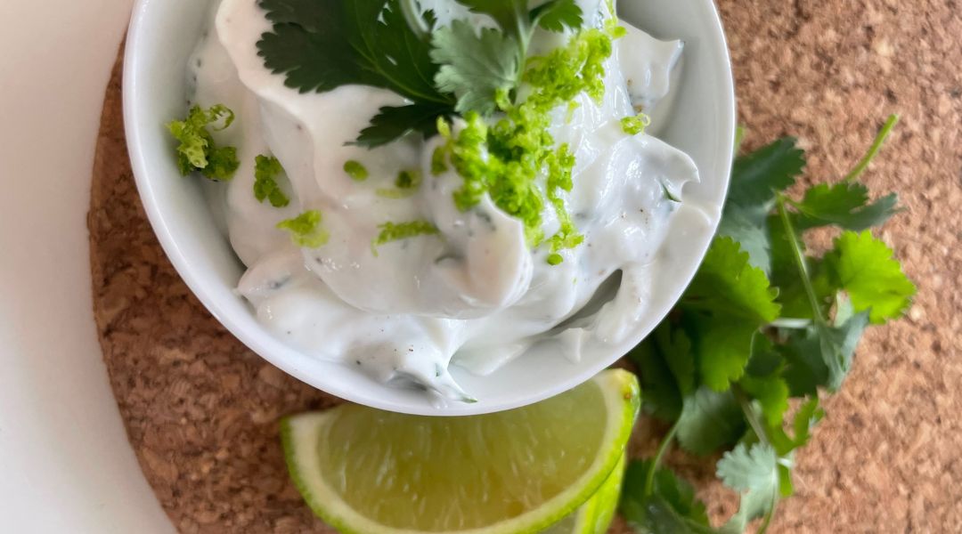 Small bowl of cilantro-lime crema with a lime wedge and fresh cilantro leaves on a cork mat