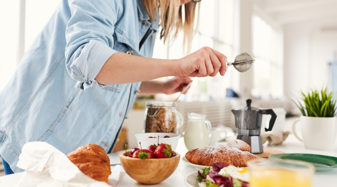 A person in a light blue denim shirt sprinkles icing sugar on a croissant at a breakfast table adorned with pastries, fruit, and coffee.