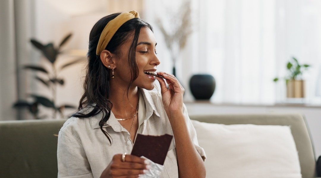 A smiling woman with a mustard-coloured headband enjoys a piece of dark chocolate while sitting on a sofa in a cozy living room.