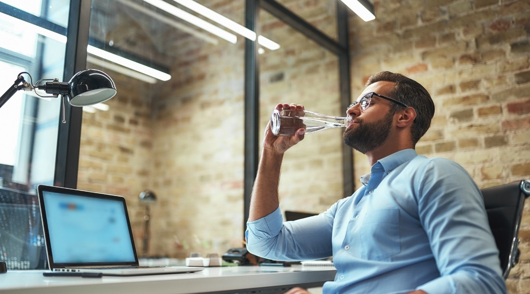 A man in a blue shirt and glasses drinks water from a glass bottle while sitting at a desk in a modern office.