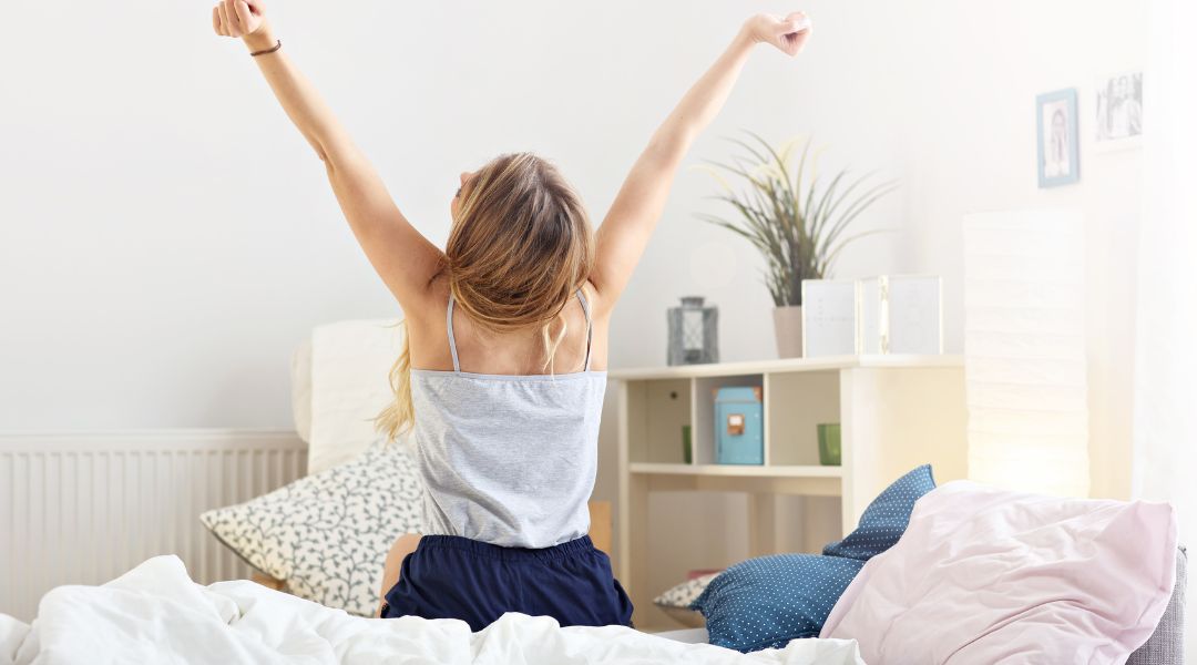 A woman stretching her arms after waking up, sitting on a bed in a bright, minimalistic bedroom with natural light