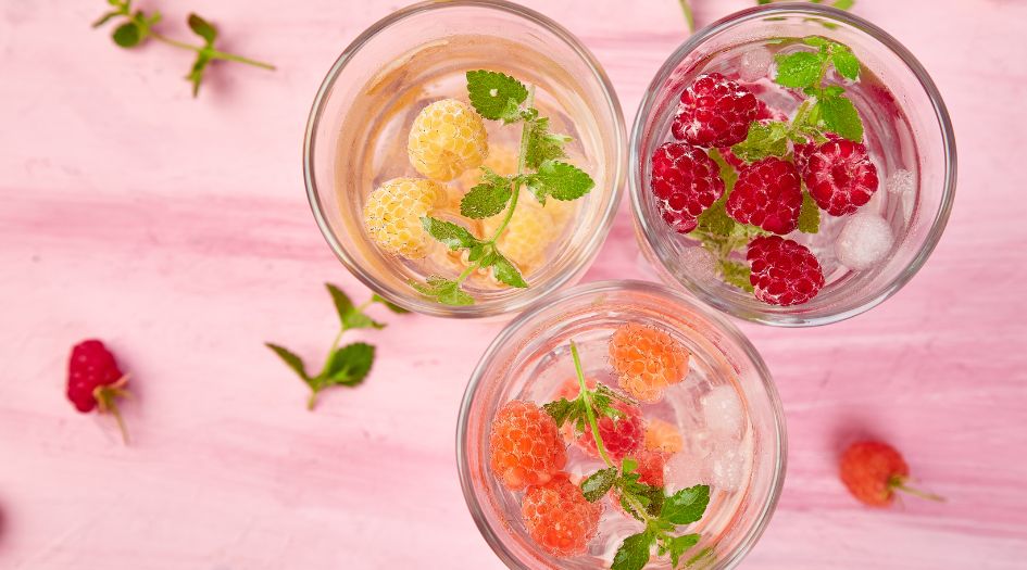 Trois verres d'eau infusée avec des framboises jaunes, rouges et orange, des feuilles de menthe et des glaçons, sur un fond rose