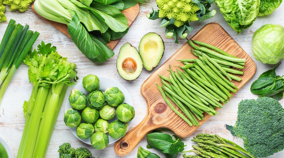 A variety of fresh green vegetables, including celery, green beans, avocado, and lettuce, arranged on a light wooden surface