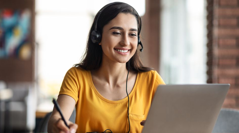 A smiling woman in a yellow shirt, wearing a headset and using a laptop, sitting in a bright indoor setting