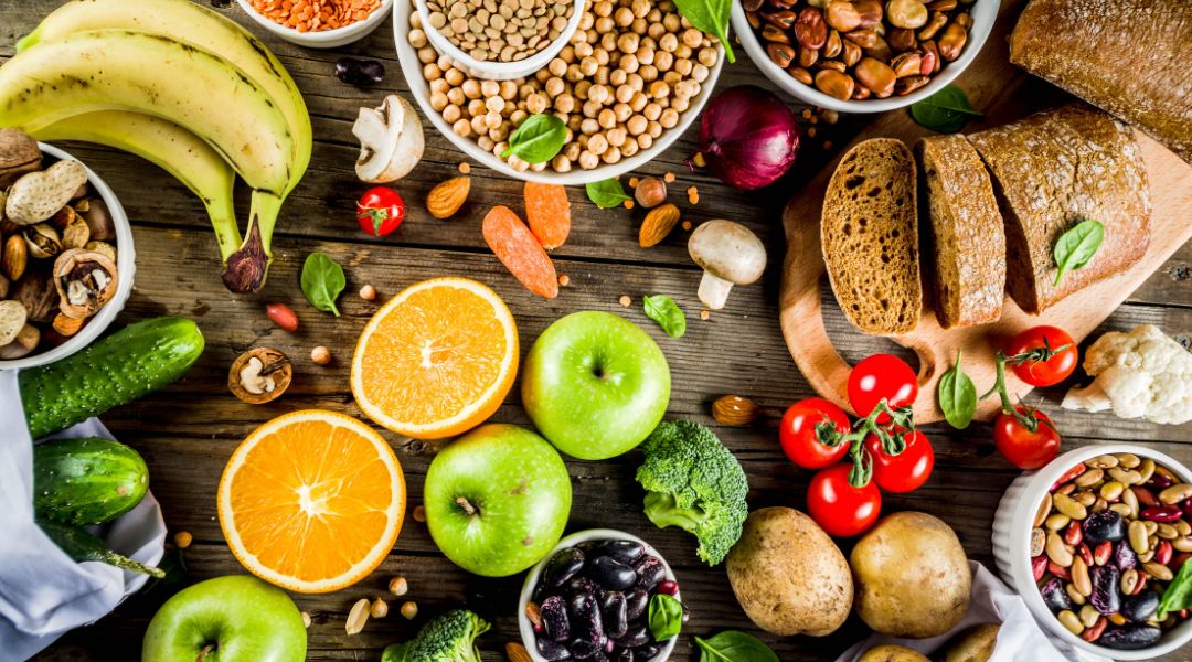 A variety of fresh fruits, vegetables, nuts, and grains, including oranges, apples, tomatoes, and bread, on a wooden surface