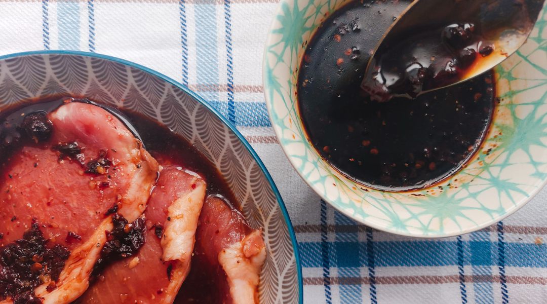 Raw pork chops marinating in a dark, spiced sauce with a spoon in a nearby bowl, placed on a blue plaid cloth