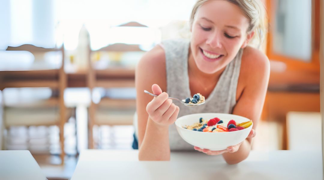 Smiling woman enjoying a bowl of oatmeal topped with fresh fruits, representing a balanced and nourishing breakfast.