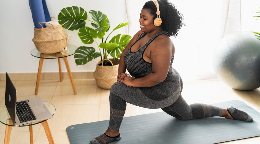 A woman performs stretches on a yoga mat in her living room, with a laptop nearby for an online session.