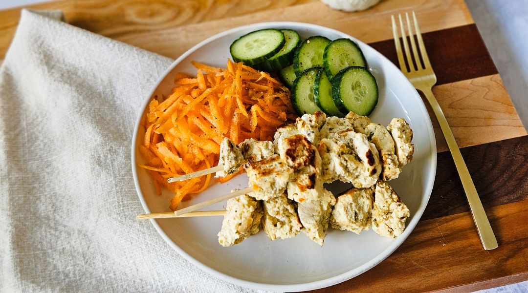 Plate of grilled tofu skewers with sliced cucumbers and shredded carrots, accompanied by a gold fork