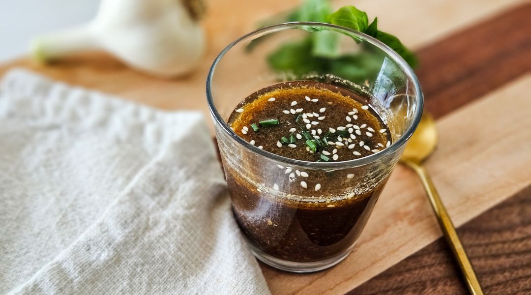 Glass of miso ginger dressing with sesame seeds and chives on top, placed on a wooden board