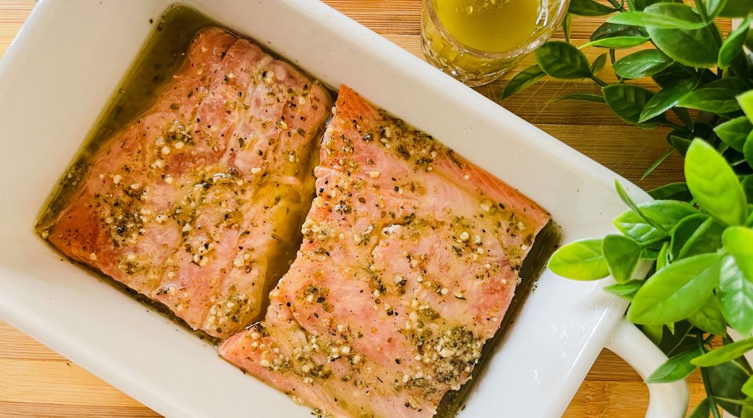 Two marinated salmon fillets in a white baking dish, topped with herbs, with a small glass of marinade nearby