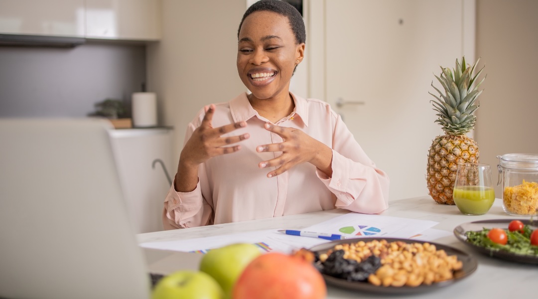 A smiling woman engages in an online nutrition consultation, surrounded by healthy foods and work documents.