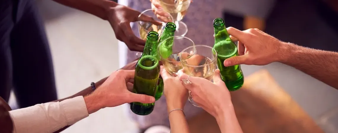 Un groupe de mains diverses trinquant avec des bouteilles de bière verte et des verres de vin dans une ambiance festive. A group of diverse hands toasting with green beer bottles and wine glasses in a celebratory setting.