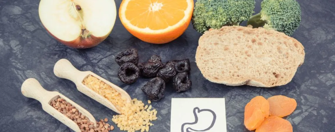 Fibre-rich foods including fruits like apples and oranges, vegetables like broccoli, whole grain bread, dried apricots, prunes, as well as cereals such as buckwheat and brown rice, placed next to an illustration of a stomach to represent potential discomforts related to the presence of FODMAPs.