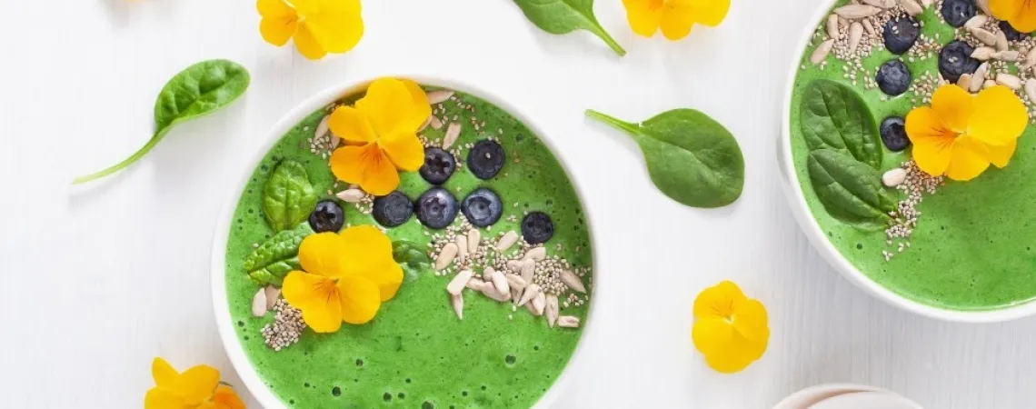 A vibrant green smoothie bowl topped with yellow edible flowers, fresh blueberries, chia seeds, sunflower seeds, and baby spinach leaves. The bowl is arranged on a light background with additional spinach leaves and yellow flowers scattered around for decoration.