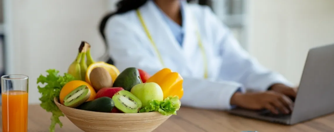 A bowl of fresh fruits, including kiwi, apple, and banana, with a glass of juice on a table, while a nutritionist works on a laptop in the background.
