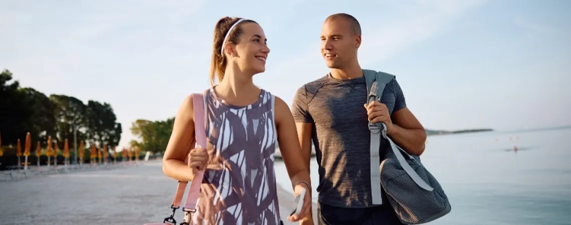 A smiling couple in activewear walks on a beach, each carrying a gym bag, with the sea and trees in the background.