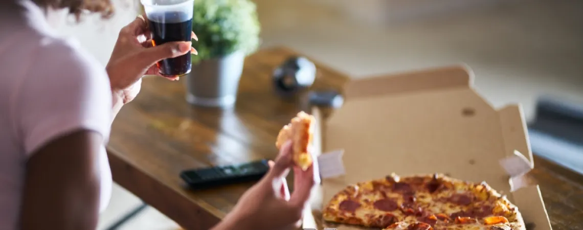 Person holding a slice of pepperoni pizza and a cup of soda, with a pizza box on a wooden table.
