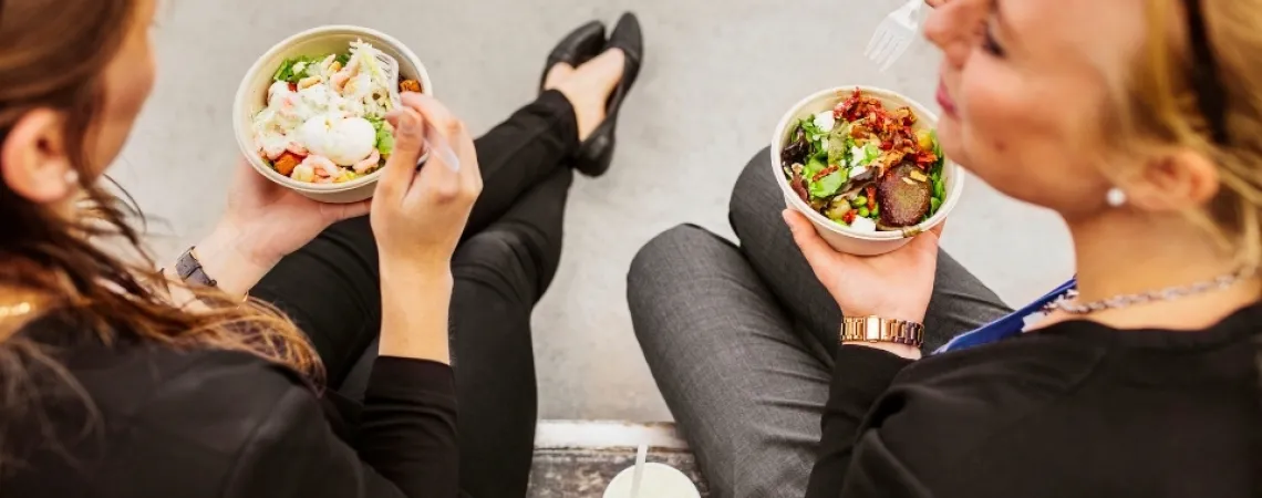 Two women seated outdoors, eating fresh salad bowls with mixed greens and toppings, accompanied by drinks.