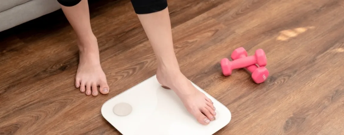 Person stepping on a white scale in a living room with pink dumbbells on the wooden floor nearby.