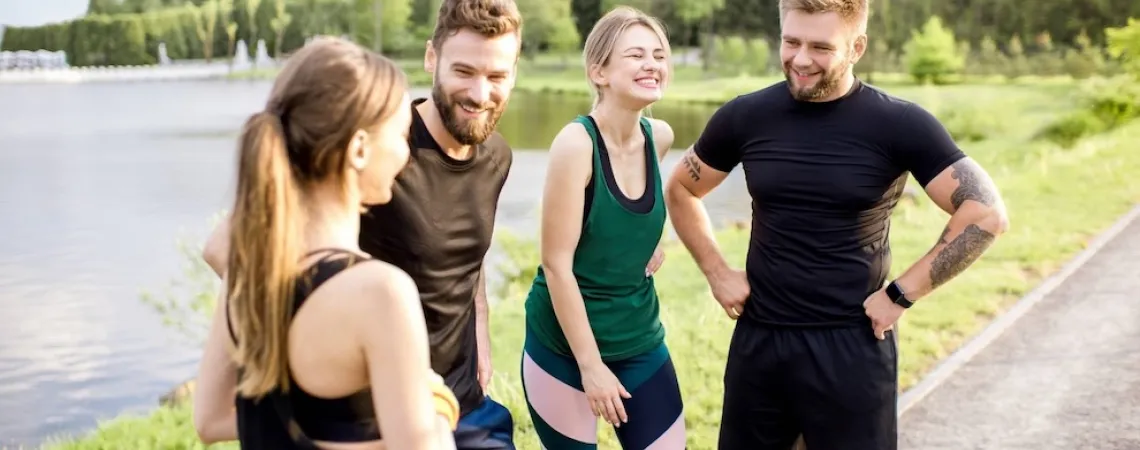 A group of four friends in activewear smiling and chatting outdoors near a lake, with a forested area in the background.