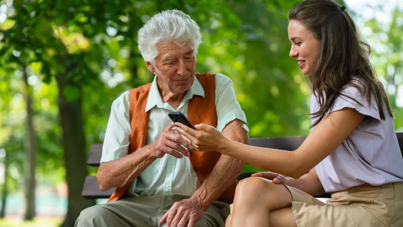 A young woman showing an elderly man how to use a smartphone while they sit on a park bench. The man has a continuous glucose monitor on his arm.