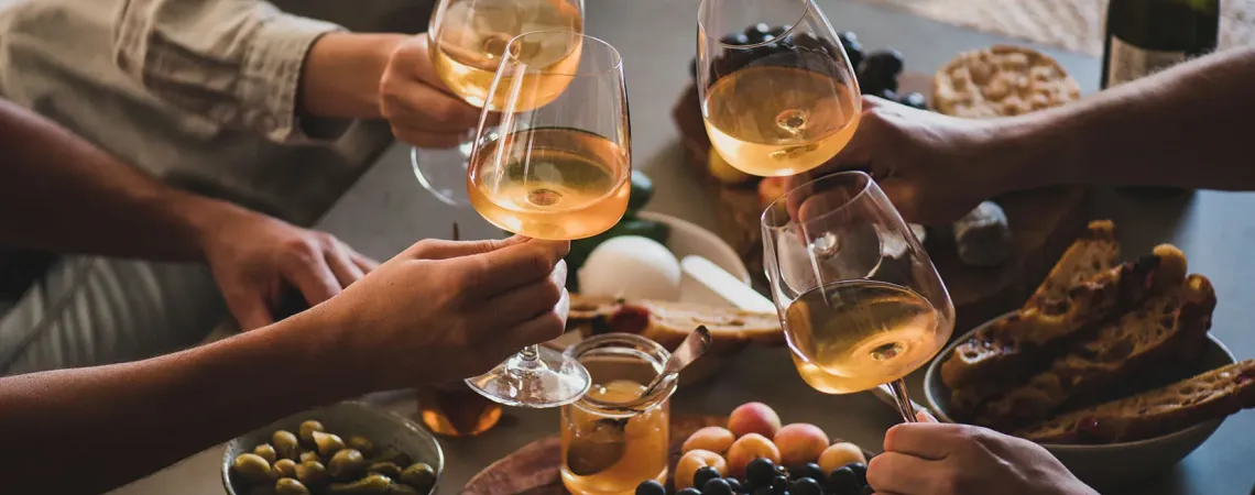 Friends toasting with white wine over a table of cheese, bread, grapes, and olives.