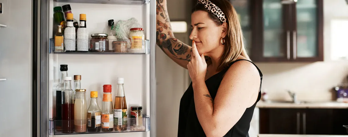 Femme debout devant un réfrigérateur ouvert, réfléchissant en regardant différents aliments et boissons à l'intérieur. Woman standing in front of an open fridge, thoughtfully looking inside at various food items and drinks.