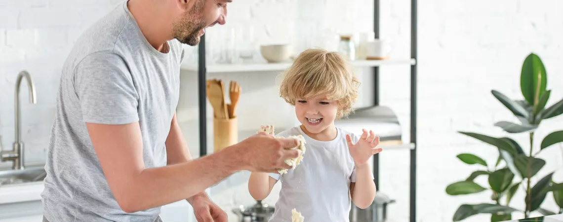 Un père et son fils rient et s'amusent en préparant de la pâte dans une cuisine moderne et lumineuse - A father and his son are laughing and having fun while making dough in a bright, modern kitchen