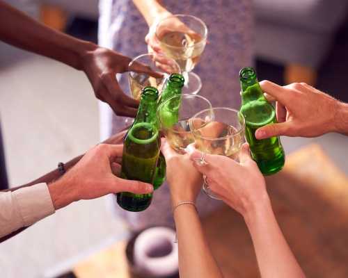 Un groupe de mains diverses trinquant avec des bouteilles de bière verte et des verres de vin dans une ambiance festive. A group of diverse hands toasting with green beer bottles and wine glasses in a celebratory setting.