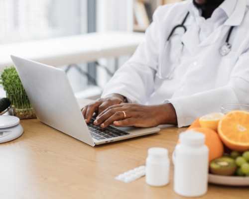 A nutritionist and Registered Dietitian in a white coat works on a laptop with fresh fruits, vegetables, and supplements on the table.