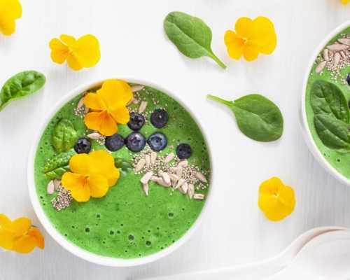 A vibrant green smoothie bowl topped with yellow edible flowers, fresh blueberries, chia seeds, sunflower seeds, and baby spinach leaves. The bowl is arranged on a light background with additional spinach leaves and yellow flowers scattered around for decoration.