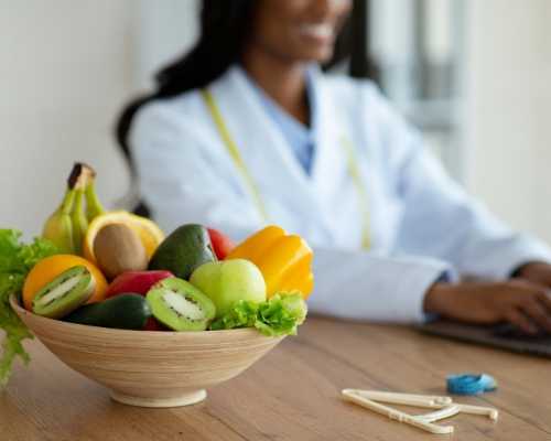 A bowl of fresh fruits, including kiwi, apple, and banana, with a glass of juice on a table, while a nutritionist works on a laptop in the background.