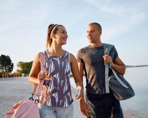 A smiling couple in activewear walks on a beach, each carrying a gym bag, with the sea and trees in the background.