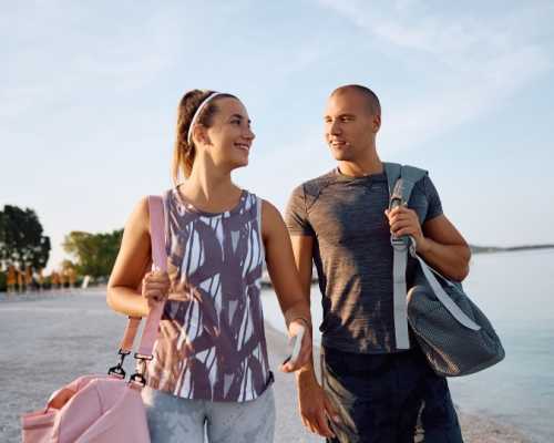 A smiling couple in activewear walks on a beach, each carrying a gym bag, with the sea and trees in the background.