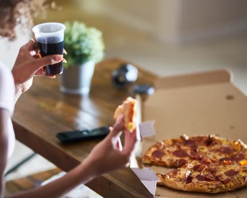 Person holding a slice of pepperoni pizza and a cup of soda, with a pizza box on a wooden table.