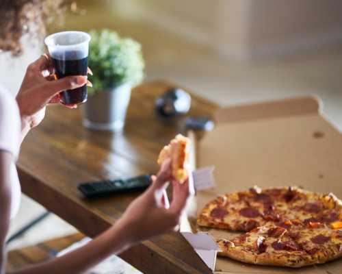 Person holding a slice of pepperoni pizza and a cup of soda, with a pizza box on a wooden table.